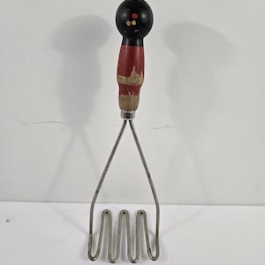 May include: A vintage potato masher with a black and red painted wooden handle and a stainless steel mashing head. The handle is round and shows signs of wear, with the red paint chipped in places. The mashing head has a wavy design.