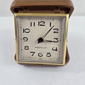 May include: A vintage Westclox travel alarm clock with a gold-tone square face and a brown protective case. The clock face is cream-colored with black numerals and hands. The clock is marked "CHINA" at the bottom.