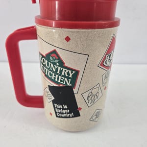 May include: A beige travel mug with a red handle and lid. The mug features a Country Kitchen logo, a black rectangular graphic with the text This Is Badger Country!, and various small illustrations of badger paw prints and diamond shapes.