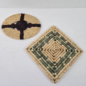 May include: Two woven trivets. One is oval with a dark brown center and cross design. The other is square with a diamond pattern in shades of green and tan. Both are made of natural fibers.