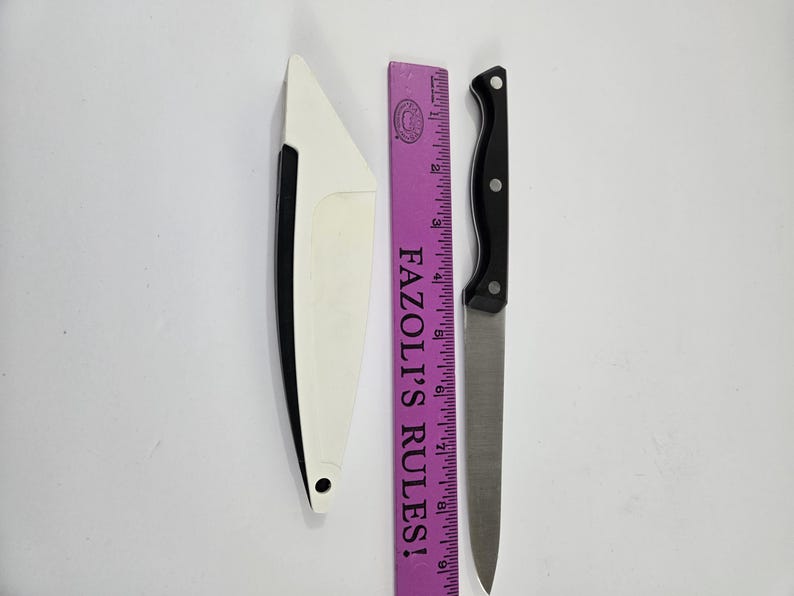 May include: A stainless steel knife with a black handle and a white knife sheath. A purple ruler with the text "FAZOLI'S RULES!" is next to the knife. The knife blade is approximately 20 cm long.