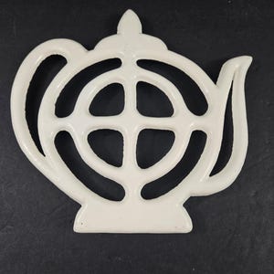 May include: White, openwork teapot-shaped trivet. The trivet is made of a solid material and has a handle, spout, and a decorative top. The design features a circular pattern in the center. The trivet is on a black surface.