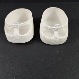 May include: A pair of white doll shoes featuring a T-strap design with molded stitching details and a decorative cutout pattern near the heel. The shoes have a rounded toe and a smooth, matte finish, presented as two separate, identical footwear items.
