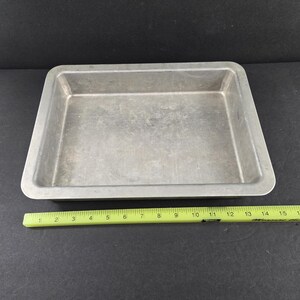 May include: A rectangular, silver-colored baking pan with slightly rounded corners. A yellow ruler is placed next to the pan, showing measurements up to 16 inches. The pan is empty and sits on a dark surface.