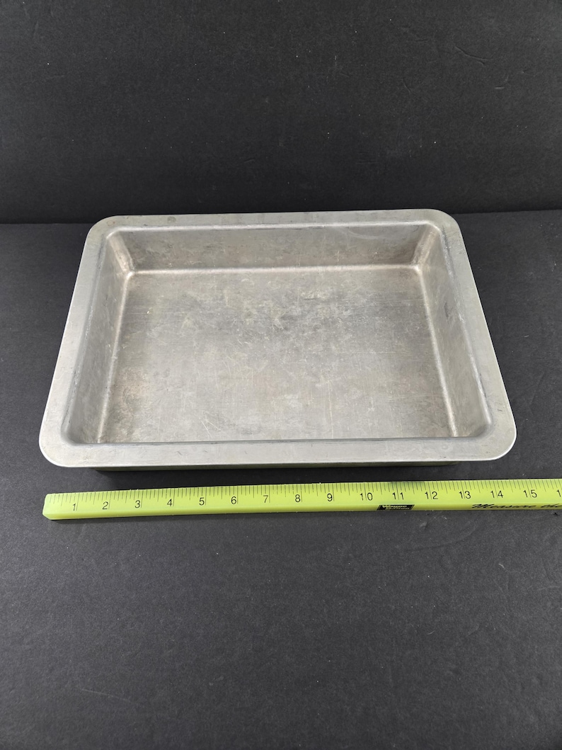 May include: A rectangular, silver-colored baking pan with slightly rounded corners. A yellow ruler is placed next to the pan, showing measurements up to 16 inches. The pan is empty and sits on a dark surface.