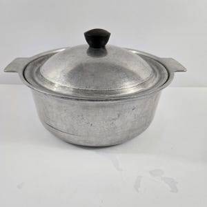 May include: A round, silver-colored aluminum cooking pot with two side handles and a matching lid. The lid features a small, black, circular knob centered on top. The pot has a smooth, metallic finish with visible signs of use and wear.