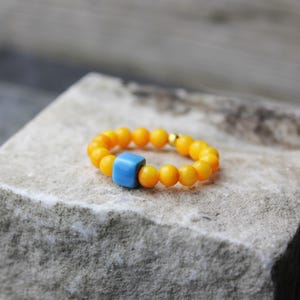 Könnte beinhalten: Ein Perlenring mit kleinen, runden, orangefarbenen Perlen und einer einzelnen, quadratischen, hellblauen Perle. Der Ring liegt auf einer Steinoberfläche. Der Ring ist ein Schmuckstück.