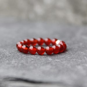 Könnte beinhalten: Ein kreisförmiger Ring, der aus kleinen, glatten, durchscheinenden tiefroten Perlen besteht. Die Perlen sind in Größe und Form einheitlich und bilden ein durchgehendes Band. Eine kleine, reflektierende Metallperle ist in die Anordnung integriert.