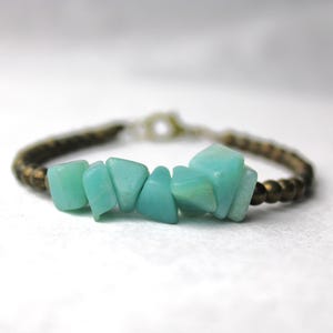 Könnte beinhalten: Ein Armband mit einer zentralen Reihe aus unregelmäßigen, polierten hellblau-grünen Steinen, flankiert von einem Band aus kleinen, dunkelmetallischen Perlen. Das Schmuckstück schließt mit einem einfachen runden Verschlussmechanismus.