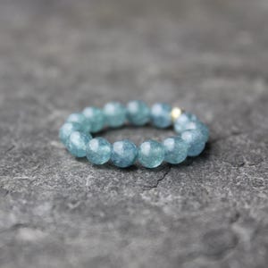 Könnte beinhalten: Ein runder Ring aus hellblauen, facettierten Perlen. Die Perlen sind zu einem durchgehenden Kreis aufgereiht. Eine kleine goldfarbene Perle ist sichtbar. Der Ring liegt vor einem dunkelgrauen, strukturierten Hintergrund.