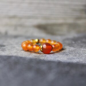 Könnte beinhalten: Ein Ring aus orangefarbenen Perlen mit einem größeren, facettierten, rot-orangen Mittelstein. Der Ring hat goldfarbene Akzente und liegt auf einer grauen, strukturierten Oberfläche. Ein Schmuckstück.
