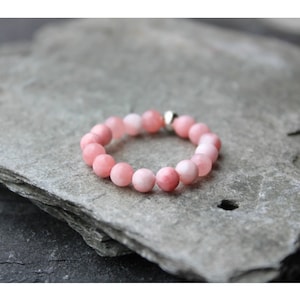 Könnte beinhalten: Ein rosa Perlenarmband mit einem silbernen Verschluss. Das Armband besteht aus runden, hellrosa Perlen. Das Armband liegt auf einer strukturierten, grauen Oberfläche.