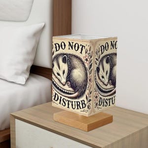 May include: A beige rectangular lamp with a wooden base. The lamp shade features a black and white illustration of an opossum curled up, with the words "DO NOT DISTURB" in a vintage font.