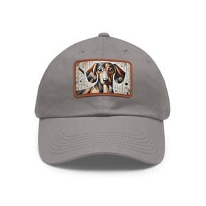 May include: A gray baseball cap featuring a patch with a geometric dachshund design. The patch has a brown border and showcases the dog's face with brown, black, and white shapes. The cap has a curved brim and a classic six-panel design.