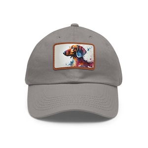 May include: A gray baseball cap featuring a watercolor painting of a dachshund dog on a rectangular patch. The dog is depicted in shades of brown, red, and blue, with a white background. The cap has a curved brim and a classic six-panel design.