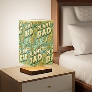May include: A green and yellow lampshade with the text "Plant Dad" repeated in a pattern. The lampshade is on a wooden base and is sitting on a wooden nightstand.