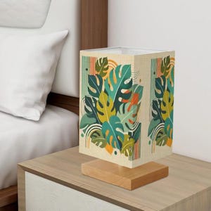 May include: A table lamp with a square linen shade featuring a colorful tropical leaf pattern. The shade is mounted on a wooden base.