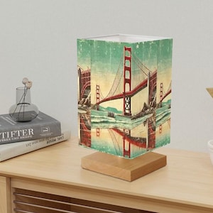 May include: A table lamp with a square shade featuring a vintage image of the Golden Gate Bridge in San Francisco, California. The shade is printed with a faded, distressed look and has a wooden base.