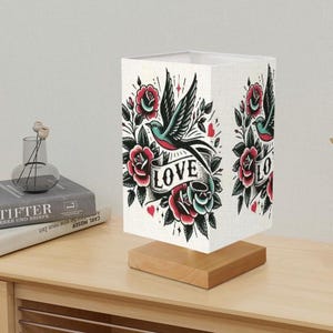 May include: A white table lamp with a square shade featuring a vintage tattoo-style design of a bird with a banner that reads "LOVE". The design includes red roses, green leaves, and hearts.