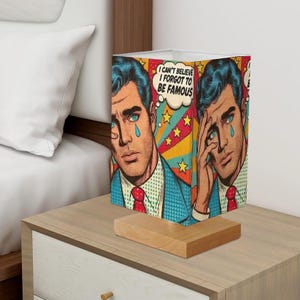 May include: A table lamp with a colorful pop art design featuring a man with a blue shirt and a red tie. The lampshade has the text "I can't believe I forgot to be famous" in a speech bubble.