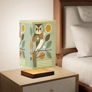 May include: A square lampshade with a geometric owl design in shades of blue, green, brown, and yellow. The lampshade sits on a wooden base.