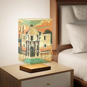 May include: A wooden table lamp with a square shade featuring a vintage illustration of the Alamo in San Antonio, Texas. The lamp is on a wooden base and is lit.