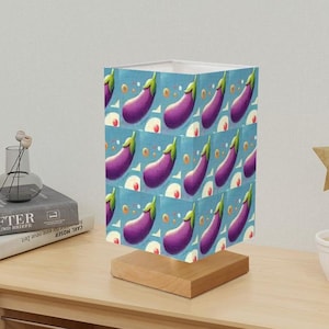 May include: A table lamp with a white shade featuring a repeating pattern of purple eggplants on a light blue background. The lamp has a wooden base.