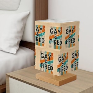 May include: A brown and beige lampshade with a repeating pattern of the text "Gay & Tired". The lampshade is on a wooden base.