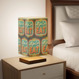 May include: A vintage-style lamp with a square shade featuring a repeating pattern of "All You Can Eat" in neon-style lettering on a teal background. The lamp is on a wooden base and sits on a wooden nightstand.