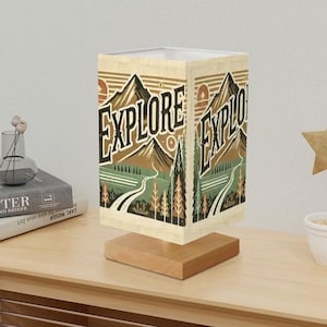 May include: A beige table lamp with a rectangular shade featuring a mountain landscape and the word "EXPLORE" printed in black. The shade is made of fabric and has a wood base.