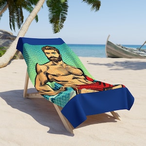 Puede incluir: Una toalla de playa azul con una ilustración de arte pop de un hombre sin camisa con barba sentado en una silla de playa. La toalla tiene un borde azul.