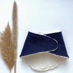 May include: A navy blue and cream-colored bib-style scarf. The outer layer is a solid navy blue fabric, while the inside is a soft, cream-colored, fuzzy material. The scarf is folded over itself.