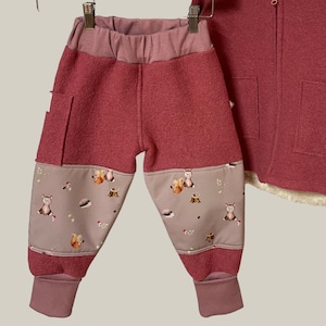 Könnte beinhalten: Eine rot-rosa Babyhose mit hellrosa Bund und Bündchen. Die unteren Beinpartien sind hellbraun mit einem sich wiederholenden Muster von Waldtieren. Eine passende rot-rosa Jacke mit Kapuze ist teilweise sichtbar.