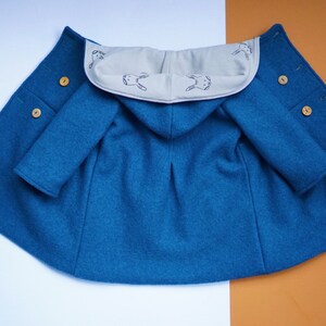 May include: A teal blue hooded cape with wooden buttons. The inside of the hood is a light beige color and features a repeating pattern of embroidered rabbits. The cape has sleeves and a rounded shape.