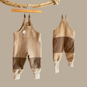 Könnte beinhalten: Zwei beige-braune Baby-Latzhosen hängen an einem Holzbügel. Die Overalls haben eine Brusttasche und braune Patches an den Knien. Die Bündchen sind gerippt und cremefarben. Ein kleines Etikett mit Logo ist sichtbar.