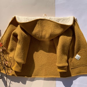 Könnte beinhalten: Senfgelbe Kapuzenjacke mit cremefarbenem Fleecefutter. Die Jacke hat einen Frontreißverschluss, eine Tasche und gerippte Bündchen. Ein kleines Etikett ist auf der Tasche aufgenäht. Die Jacke ist offen und zeigt das Innenfutter und die Ärmel.