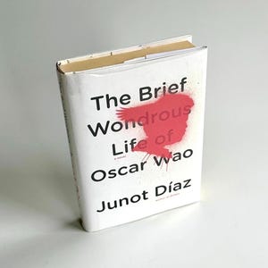 Puede incluir: Un libro de tapa dura titulado "La breve y maravillosa vida de Oscar Wao" de Junot Díaz. El libro tiene una cubierta blanca con texto negro y un gráfico rojo. El libro está ligeramente inclinado.