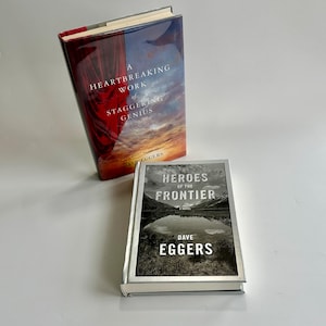 Puede incluir: Dos libros de tapa dura. El libro superior tiene una cubierta roja y azul con el título "A Heartbreaking Work of Staggering Genius" de Dave Eggers. El libro inferior se titula "Heroes of the Frontier" de Dave Eggers, con una imagen de paisaje.