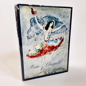 May include: A book cover featuring a watercolor painting of a ballerina in a red, blue, and white dress, with a red bird flying above her. The text "Marc Chagall "The Ballet"" is printed in black at the bottom of the cover.