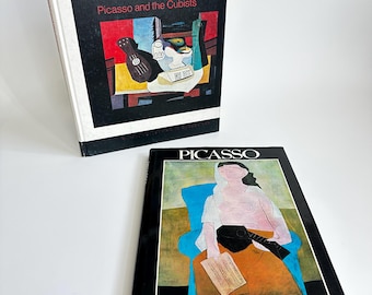 May include: Two books about Pablo Picasso. The first book is titled "Picasso and the Cubists" and features a painting of a still life with a guitar, a bottle, and a glass. The second book is titled "PICASSO" and features a painting of a woman sitting in a chair holding a guitar.