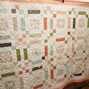 May include: A white quilt with a floral and geometric pattern. The quilt features squares and rectangles in shades of peach, green, and brown. The quilt has a scalloped edge and is likely handmade.