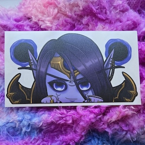 以下が含まれることがあります： 長くて濃い紫色の髪と尖った耳を持つ、青い肌のキャラクターが描かれた長方形のステッカー。キャラクターは大きな青い目を持ち、金と黒のアクセントで飾られています。ステッカーは、ふわふわのマルチカラーの背景に設定されています。