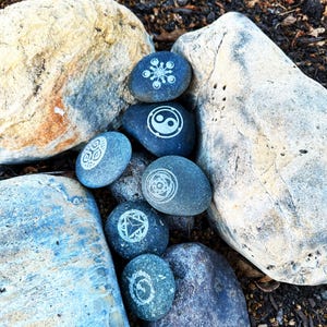 May include: A collection of seven smooth, gray stones with white painted symbols, including a yin yang, a snowflake, and a geometric design. The stones are arranged on a bed of brown dirt and rocks.