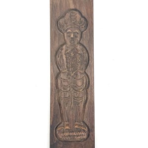 May include: A rectangular, dark brown wooden cookie mold featuring a carved image of a chef. The chef is depicted wearing a tall hat, a jacket with buttons, and pants. The mold is designed for creating detailed cookies.