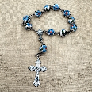 May include: A rosary with white beads decorated with blue floral designs. The rosary features a silver-toned crucifix and a chalice-shaped centerpiece. The beads are strung together with a silver-colored chain.