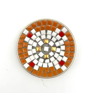 May include: A round mosaic coaster with a silver metal rim. The mosaic design features a pattern of orange, white, and red square tiles. The center has gold-colored square tiles.