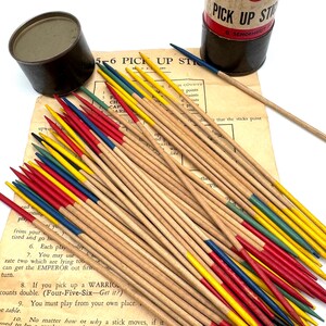 May include: A vintage Pick Up Sticks game set. The game includes a cylindrical container, a small tin, and a set of wooden sticks with colored tips in red, yellow, blue, and green. The game instructions are visible in the background.