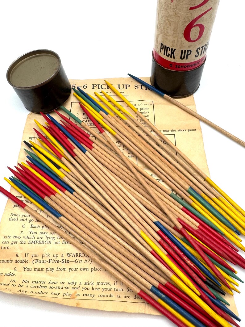 May include: A vintage Pick Up Sticks game set. The game includes a cylindrical container, a small tin, and a set of wooden sticks with colored tips in red, yellow, blue, and green. The game instructions are visible in the background.