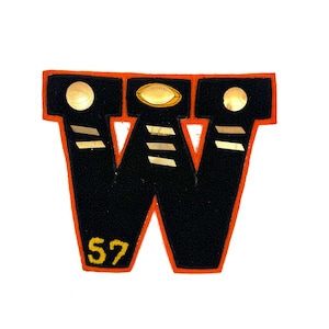May include: A vintage black felt patch in the shape of the letter "W" with an orange border. The patch features gold accents, including two circles and an oval, and the number "57" in yellow.
