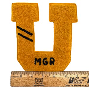 May include: Mustard yellow felt letter "U" patch with black diagonal stripes and the letters "MGR". A wooden ruler is below the patch. The ruler has text that reads "MADE IN USA" and "Make It A RULE To Call 644-4404".
